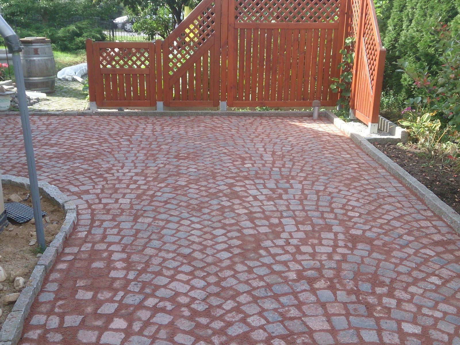 Hoffläche aus rotem Porphyr-Kleinpflaster im Segmentbogenverband mit eingestreuten grauen Granitsteinen, eingefasst von Granitbordsteinen, dahinter ein roter Holzsichtschutz
