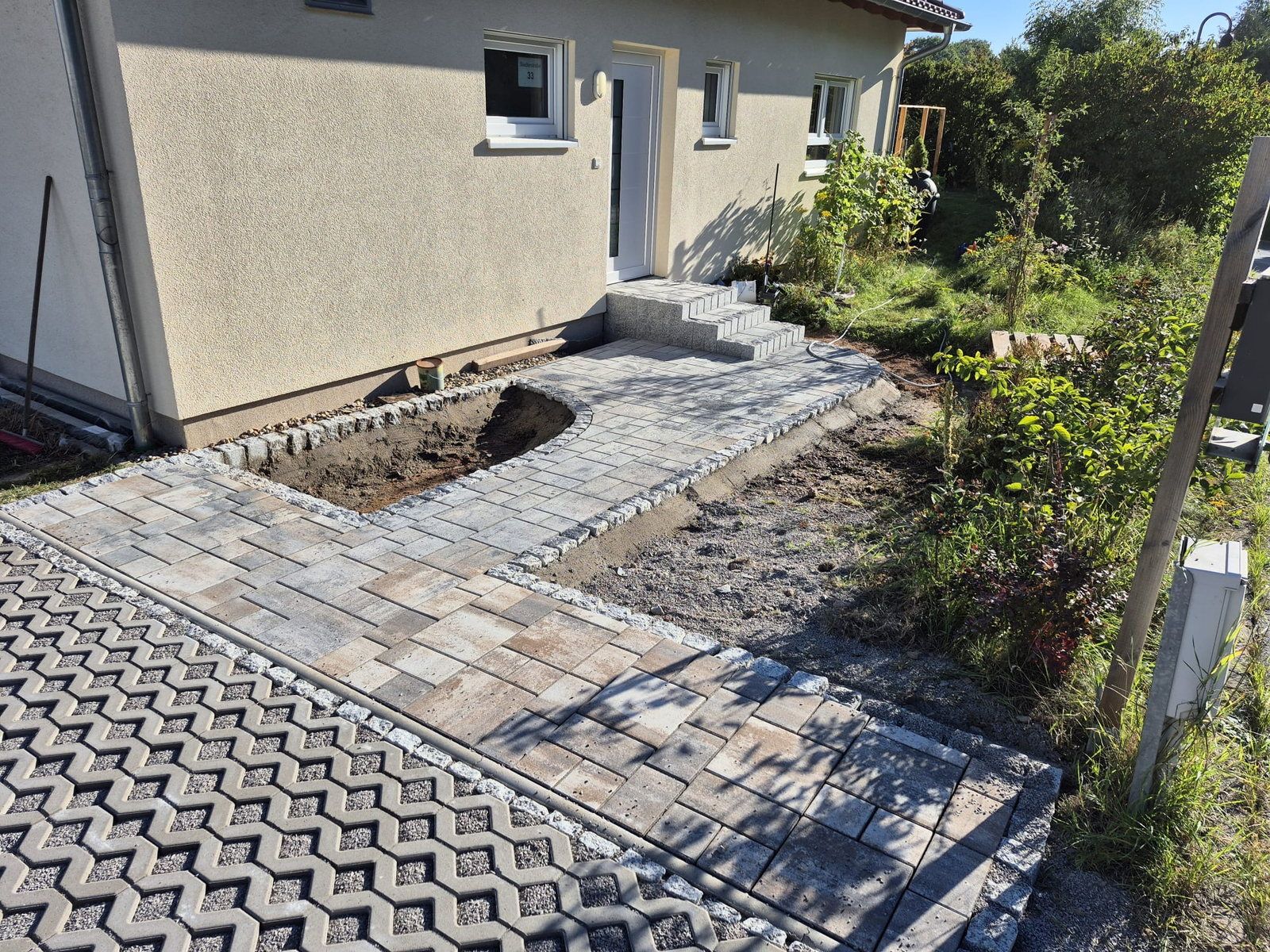 Neu verlegter Plattenweg mit Granitrandsteinen entlang einer Hausfassade, eine rechteckige Fläche ist für ein Pflanzbeet ausgespart, im Vordergrund ein Ökopflaster-Stellplatz