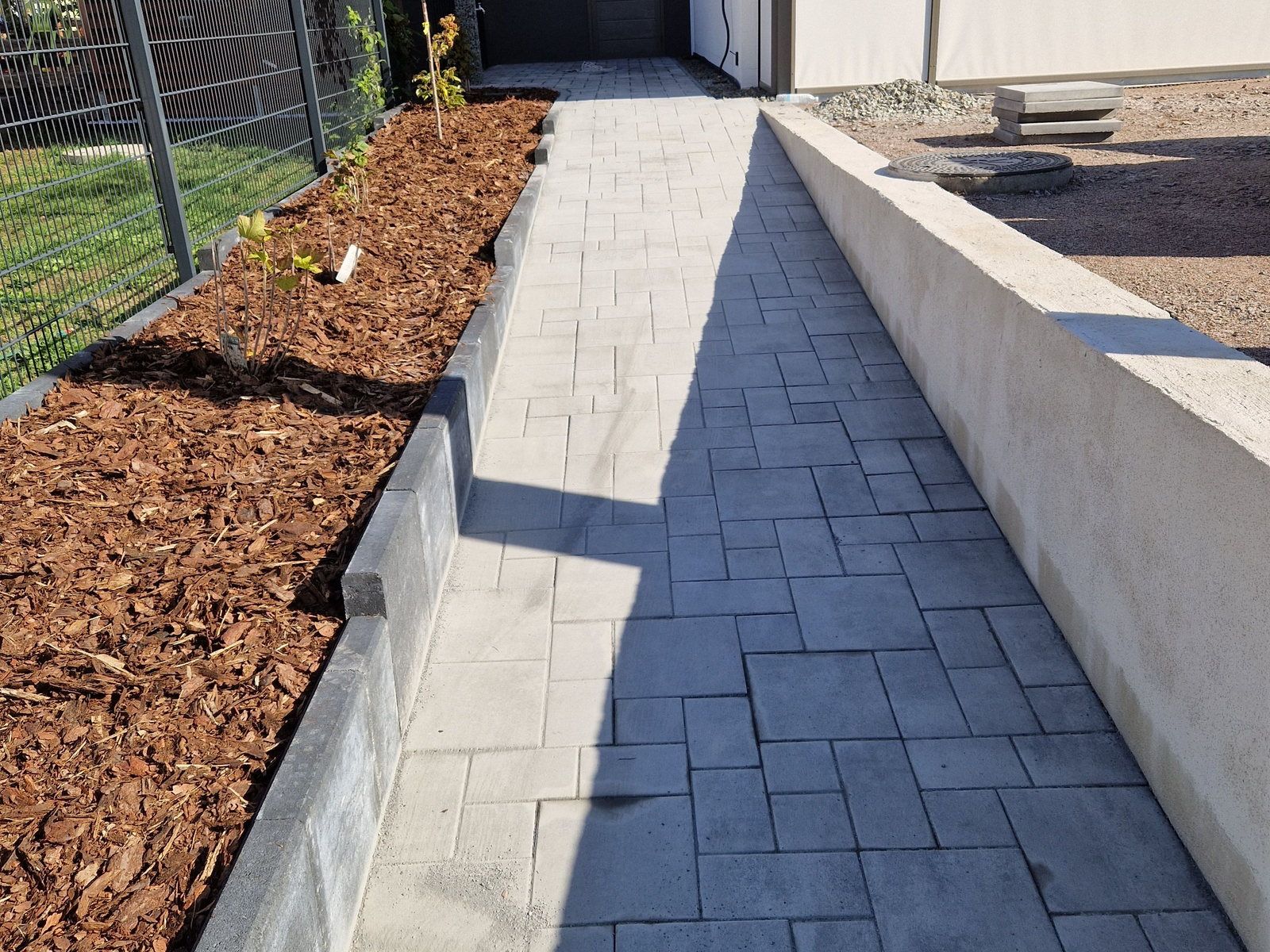 Anthrazitfarbener Plattenweg entlang einer Hauswand, links eine niedrige Stützmauer aus Betonwinkelelementen mit Rindenmulchbeet und jungen Pflanzen, rechts eine helle Mauer