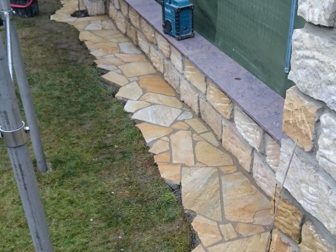 Sandsteinmauerwerk mit heller Abdeckplatte, davor ein Streifen aus verlegten Polygonalplatten entlang der Rasenkante, im Hintergrund eine Baustellenabhängung