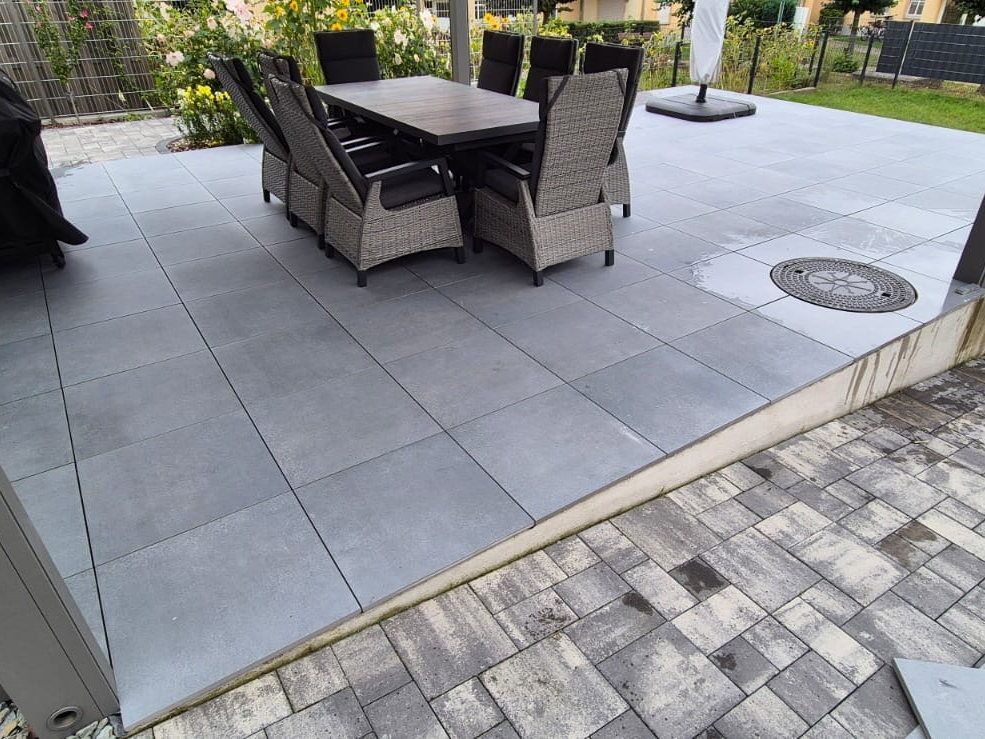 Terrasse aus großformatigen grauen Fliesen mit Sitzgruppe unter einer Pergola, im Vordergrund ein Bodenablauf, rechts angrenzend eine Fläche aus rechteckigem Betonpflaster