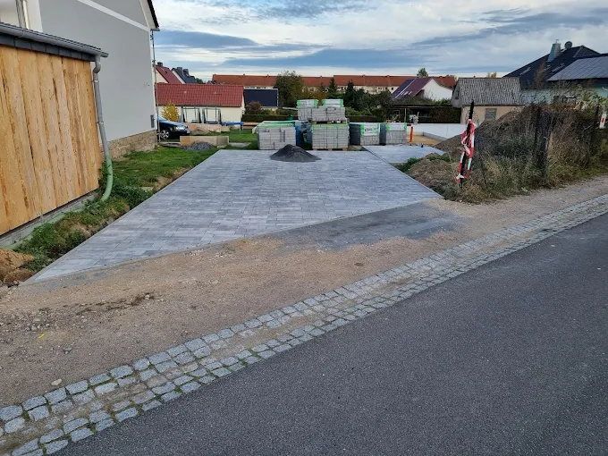 Neu gepflasterte Hofzufahrt aus grauem Betonsteinpflaster, zur Straße hin abgeschlossen durch ein breites Band aus Granit-Kleinpflaster, seitlich Paletten mit Pflastermaterial