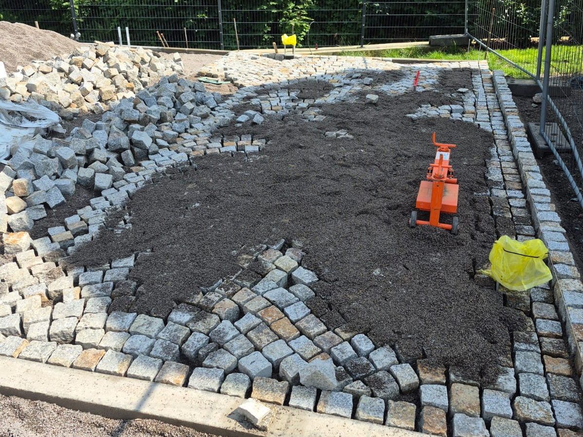 Runde Pflasterfläche aus Natursteinpflaster mit ausgesparter kreisrunder Fläche für ein Beet in der Mitte, Rüttelplatte und Bauzaun im Hintergrund