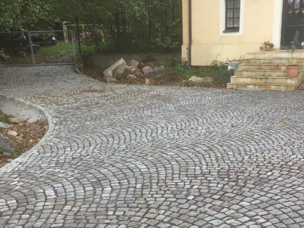 Geschwungene Hofeinfahrt aus Natursteinpflaster im Fächerverband vor einem hellgelben Haus, mit Gartentor, Natursteinmauer und Freitreppe zur Haustür