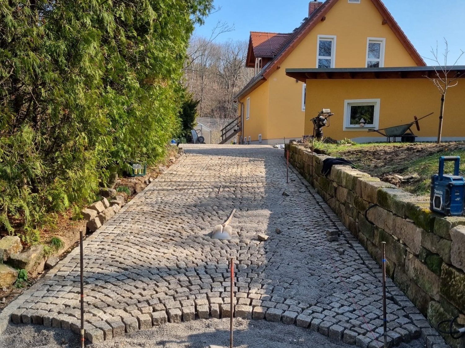 Kopfsteinpflaster-Auffahrt aus Granit im Fächerverband entlang einer Natursteinmauer, im Hintergrund ein gelbes Haus, links eine hohe Thuja-Hecke