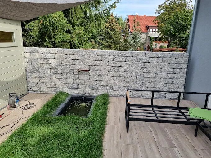 Terrasse mit hellen Dielen in Holzoptik, davor ein schmales längliches Wasserbecken in einem Rasenstreifen, dahinter eine Mauer aus grauen Granit-Mauersteinen