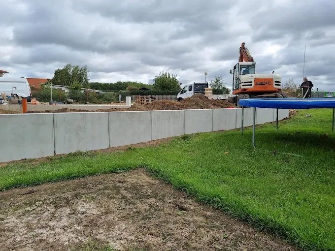 Lange Stützmauer aus Betonwinkelelementen an einer Grundstücksgrenze, davor eine angelegte Rasenfläche, im Hintergrund ein Kettenbagger und ein Transporter